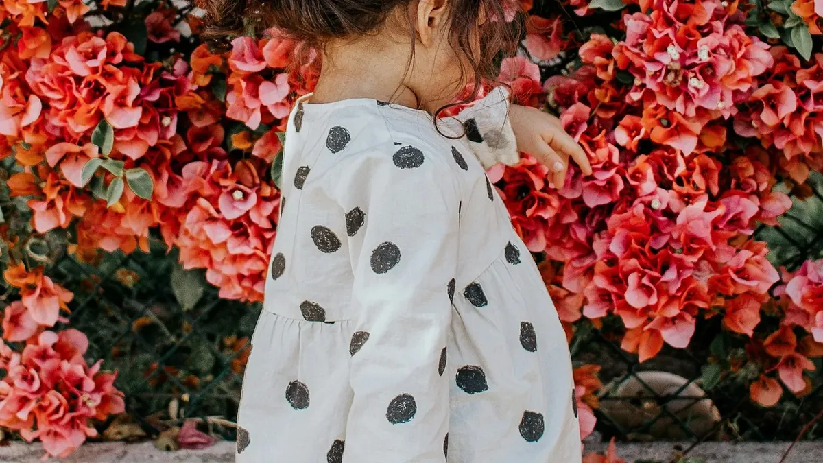 Girl in black and white polka-dot long-sleeved blouse and blue shorts standing beside red petaled flowers during daytime