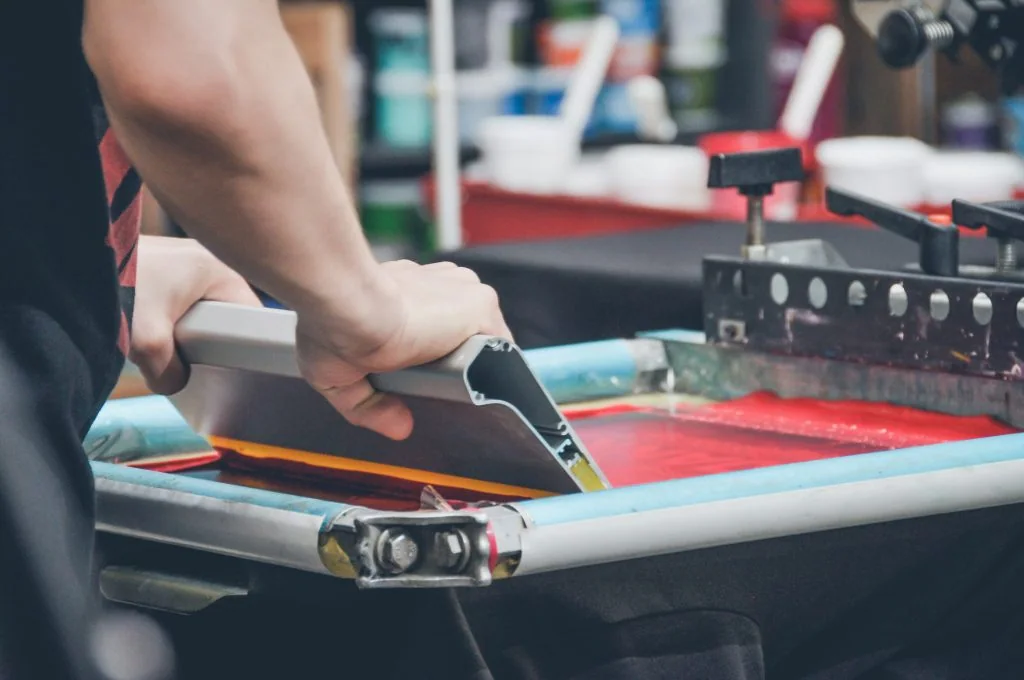 A print operator pulling a squeegee across a screen coated in red ink.