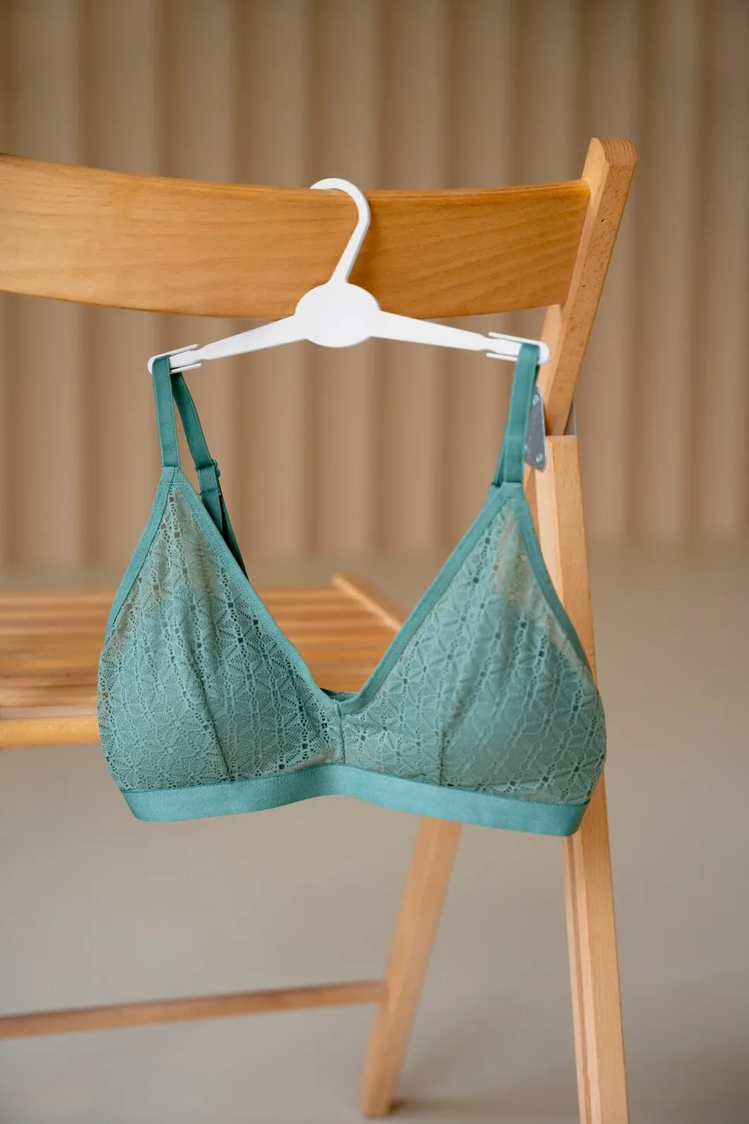 Une bralette en dentelle bleu canard, à bretelles réglables, suspendue à une chaise en bois.