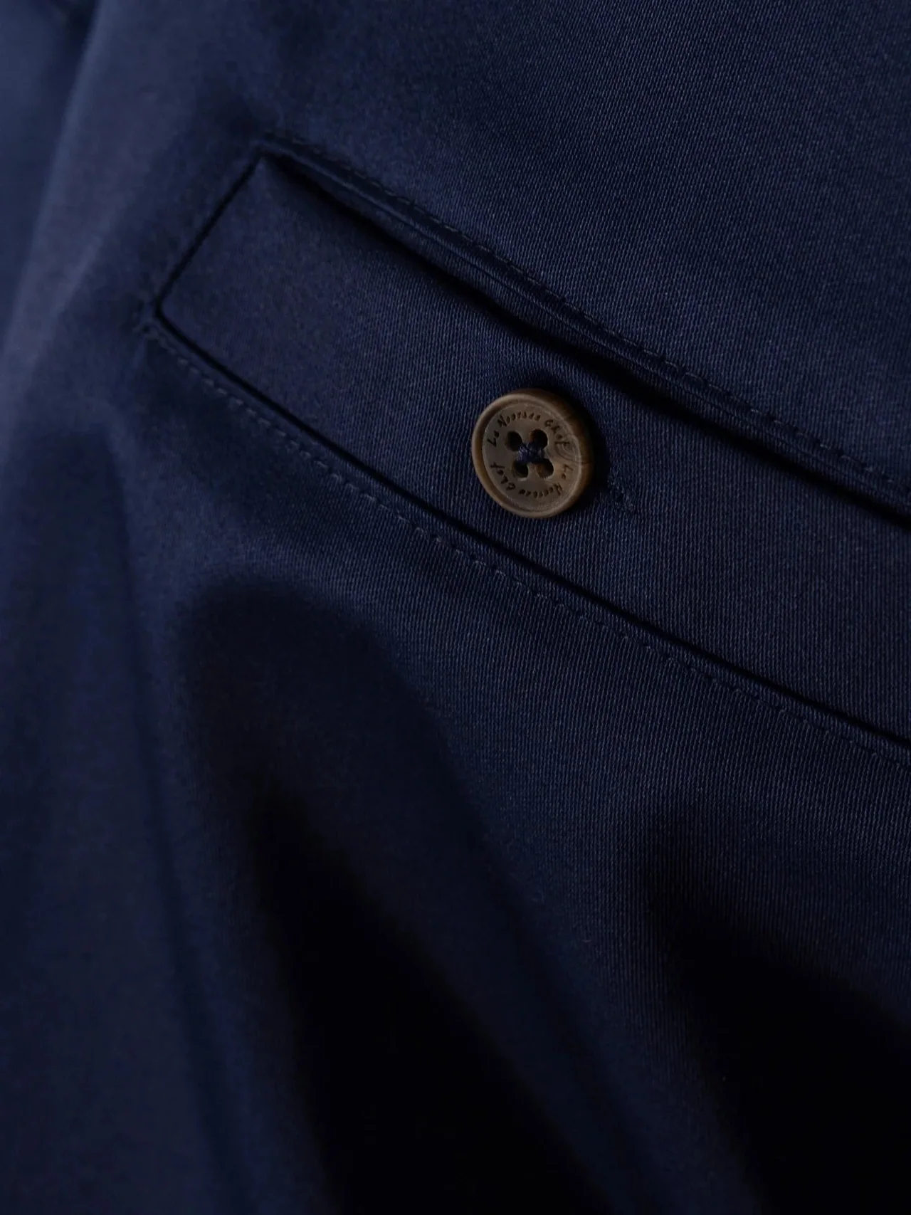 Close view of a navy chef jacket pocket showing the button, pocket opening and edge stitching.