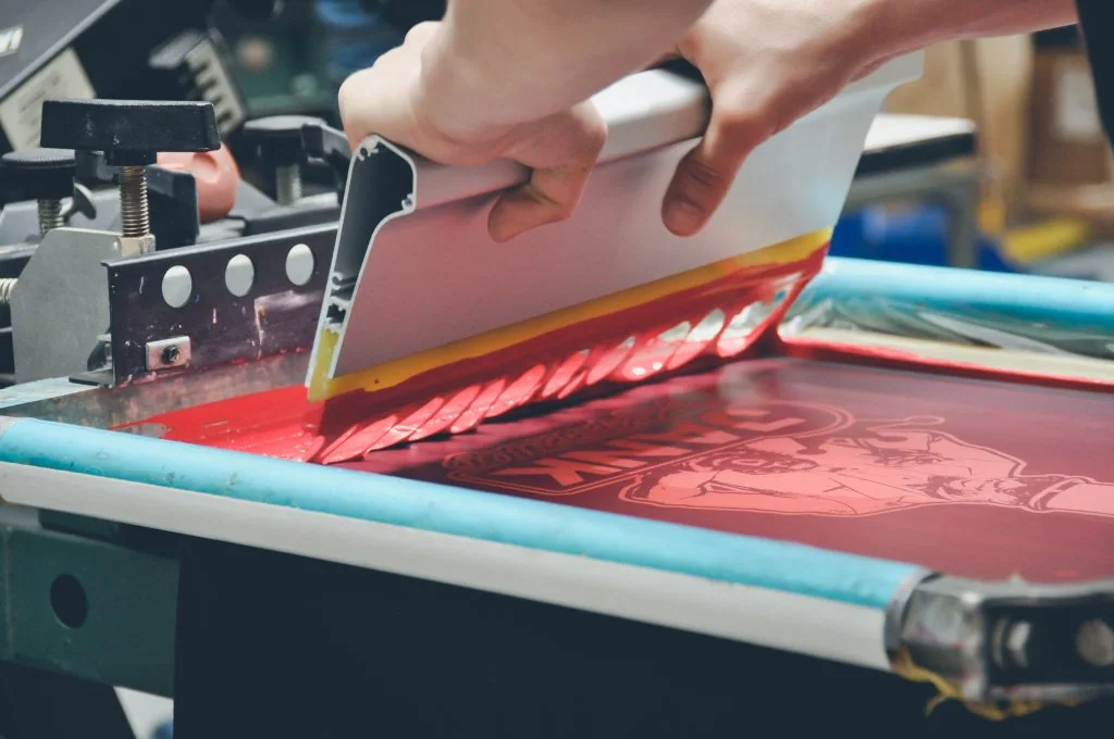 Close view of a squeegee lifting from red ink on a garment printing screen.