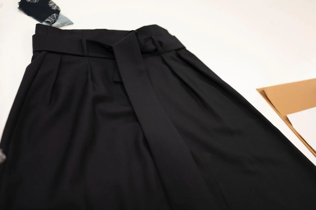 Close view of black trouser pleats, waistband and pocket opening on a worktable.