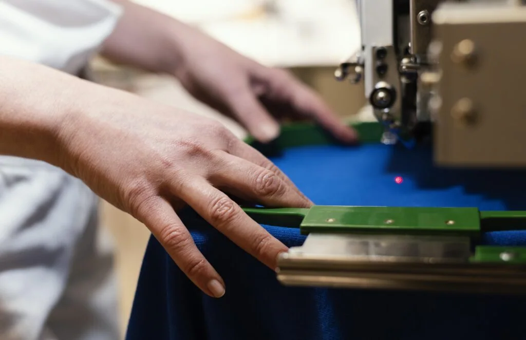Hands guiding blue fabric beneath an industrial machine needle.