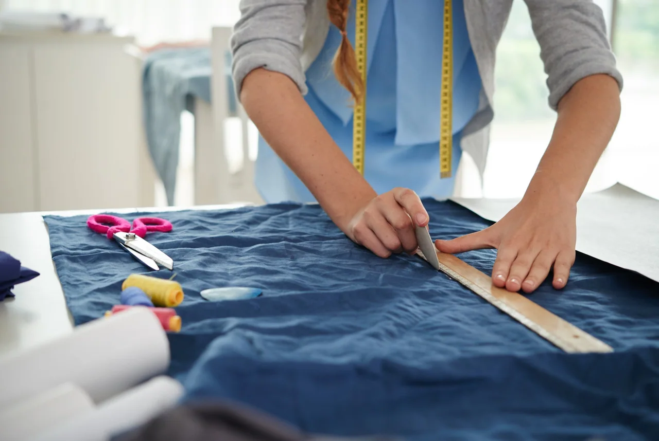 Hands marking blue fabric with a ruler on a cutting table.