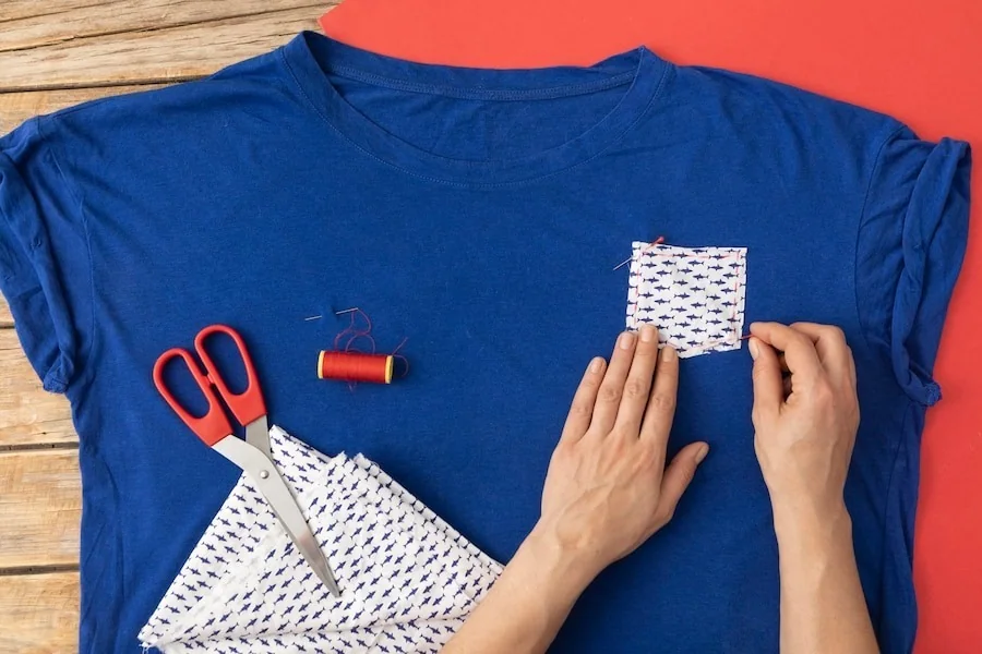 Hands positioning a patterned chest pocket on a blue T-shirt beside sewing tools.