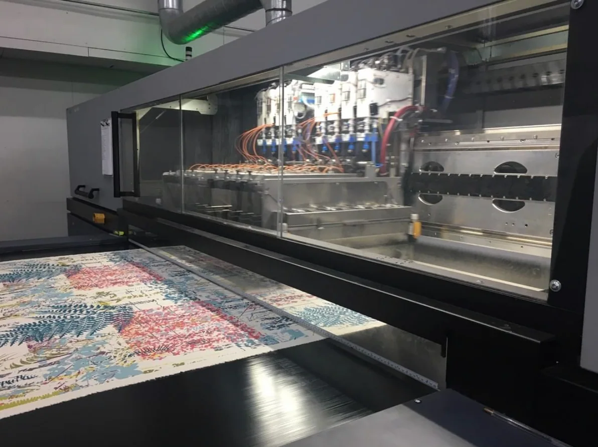 Patterned fabric emerging from an industrial textile printer with its printhead assembly visible.