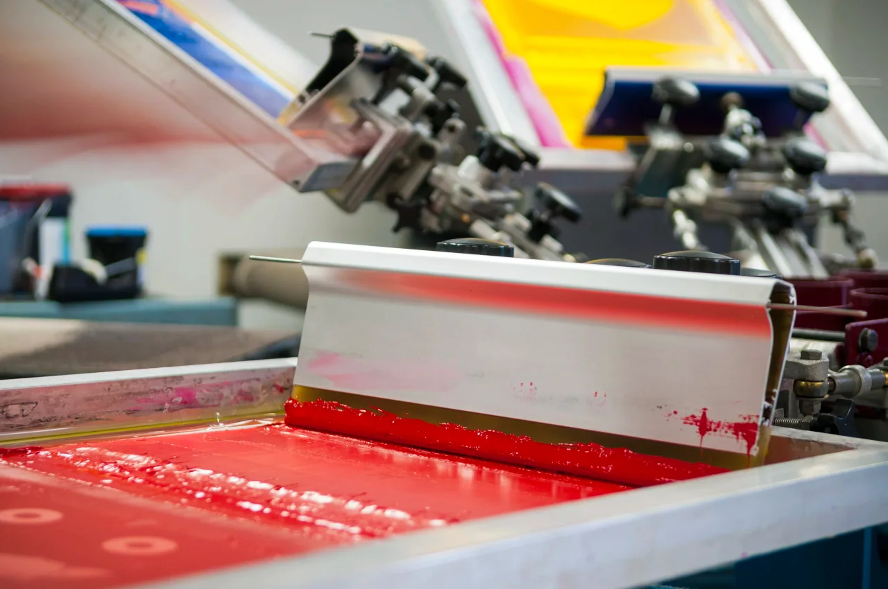 Red screen-printing ink spread across a mesh screen beneath a metal squeegee.